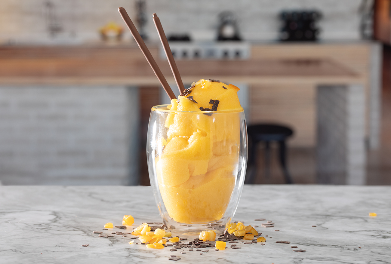 Ein Glas mit cremigem, gelb leuchtendem veganem Mango-Eis steht auf einer Küchenarbeitsplatte. Zwei Schokoladenstäbchen stecken im Eis, einige kleine Mangostückchen und Schokosplitter liegen daneben. Das Dessert veranschaulicht ein selbstgemachtes veganes Eis.