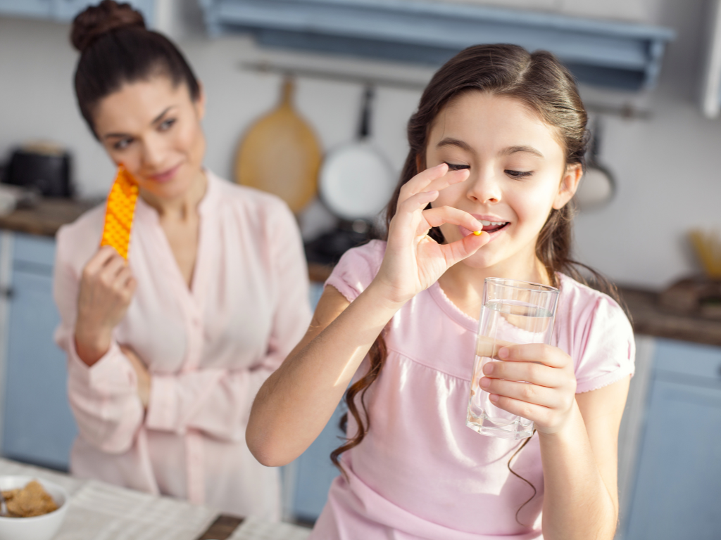 Mädchen nimmt eine Vitamintablette ein und hält ein Glas Wasser in der Hand, Mutter ist lächelnd im Hintergrund