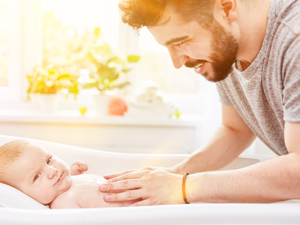 Foto eines Babys in einer Badewanne, Vater streichelt liebevoll über den Bauch des Säuglings